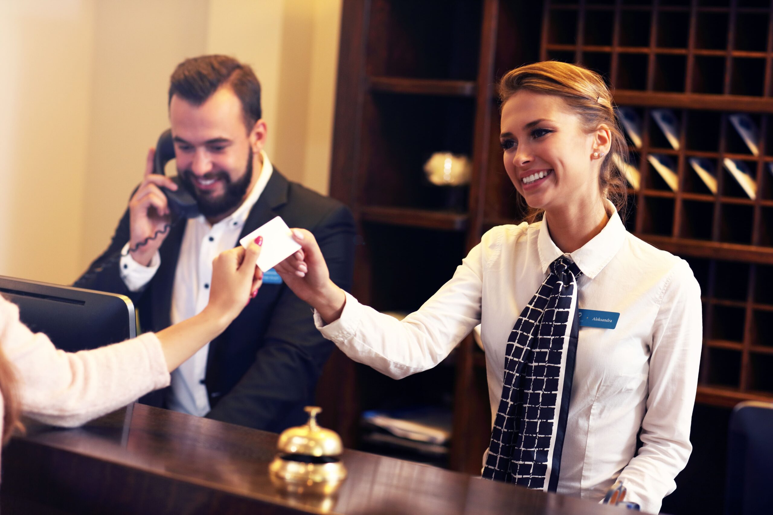 Hotel receptionist handing a key card to a guest at the front desk while another staff member speaks on the phone.
