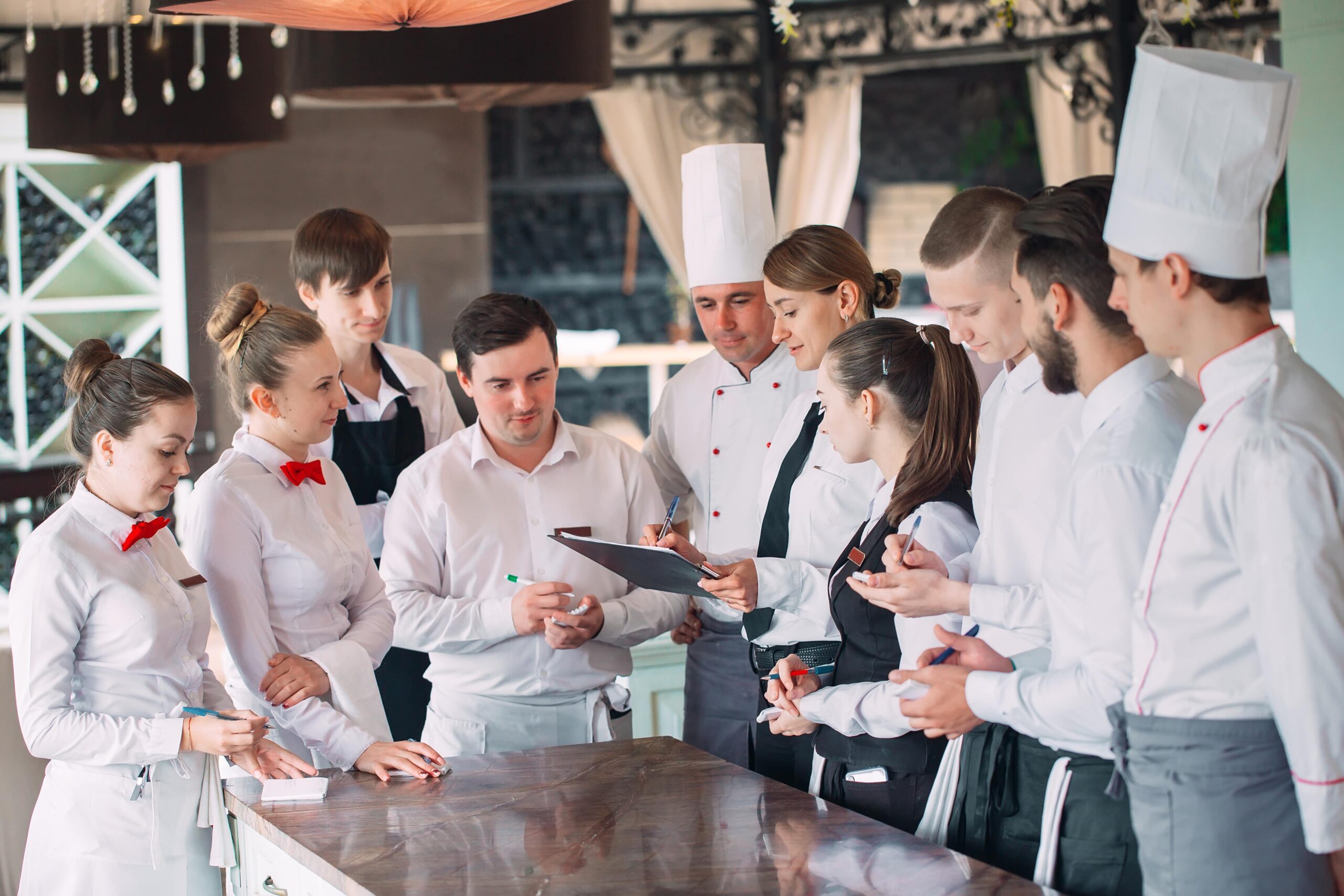Restaurant team gathered around a counter for a briefing, with chefs in white hats and servers in uniform holding notepads.